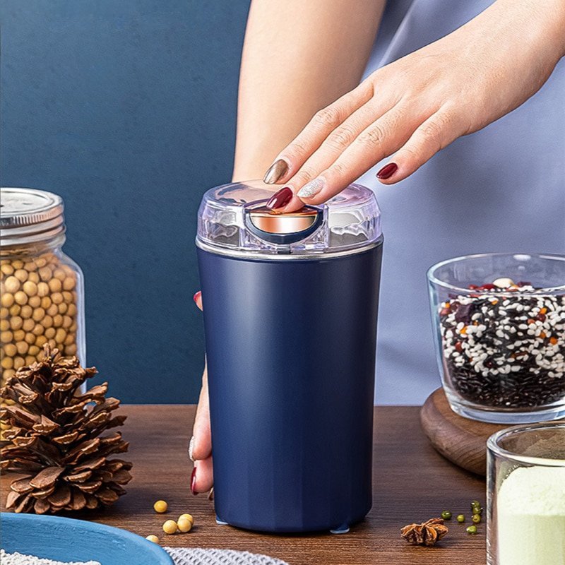 Elektrische Kaffeemühle in Dunkelblau auf Holztisch, umgeben von verschiedenen Bohnen, Getreide, Sojabohnen und pürierter lila Paste