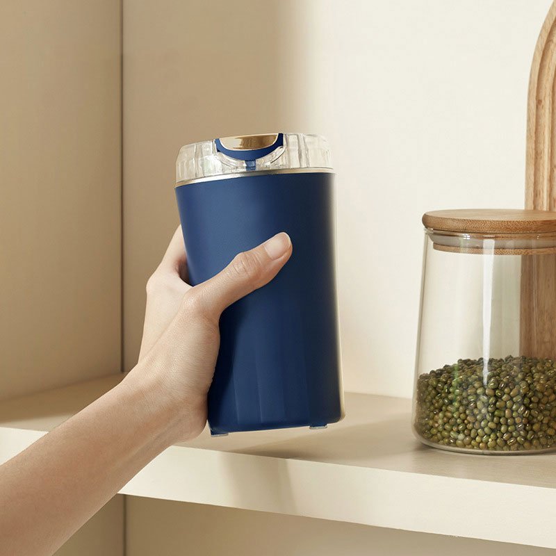 Elektrische Kaffeemühle in Dunkelblau auf Holztisch, umgeben von verschiedenen Bohnen, Getreide, Sojabohnen und pürierter lila Paste
