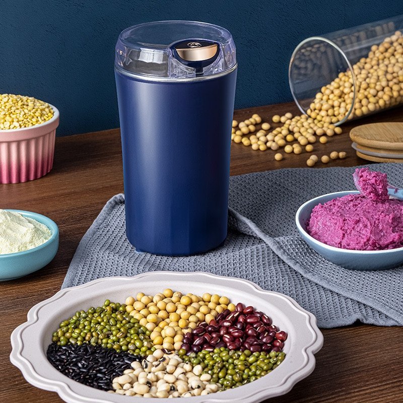 Elektrische Kaffeemühle in Dunkelblau auf Holztisch, umgeben von verschiedenen Bohnen, Getreide, Sojabohnen und pürierter lila Paste