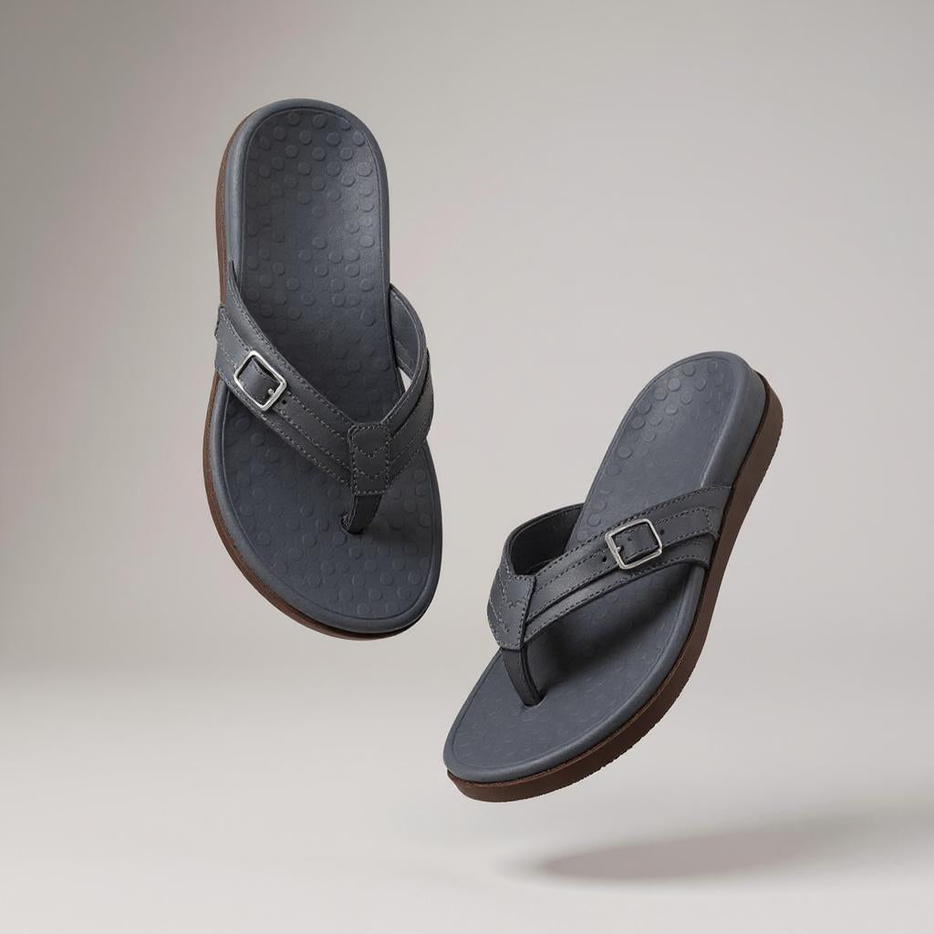 Ein Paar rosafarbene Zehensandalen mit Schnalle, beigefarbenem Fußbett und brauner Sohle, schwebend vor grauem Hintergrund.